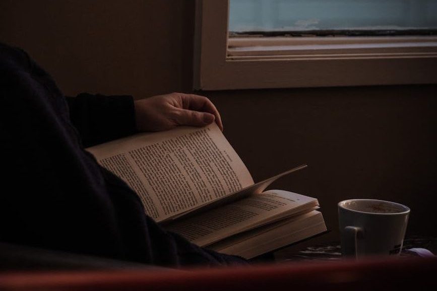 A person reading a book by a window with a coffee cup nearby, creating a quiet and reflective atmosphere.