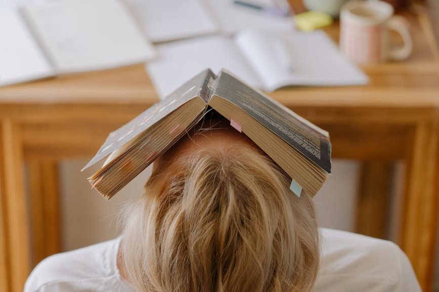 Frustrated reader with an open book covering their head, sitting at a cluttered desk during a reading slump.